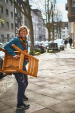 Lachende Frau geht mit Stuhl unterm Arm auf Bürgersteig. Foto: Katja Schneider